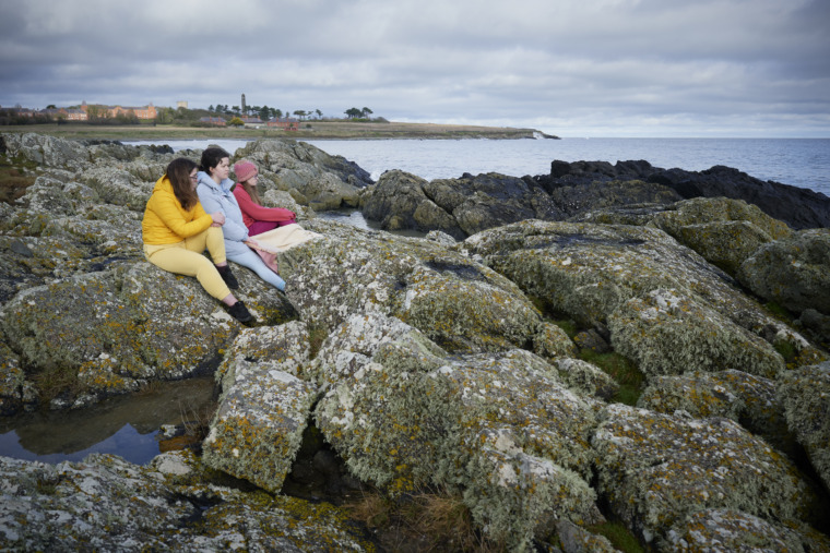 Photo of three young people sitting on a coastal rocky outcrop.
